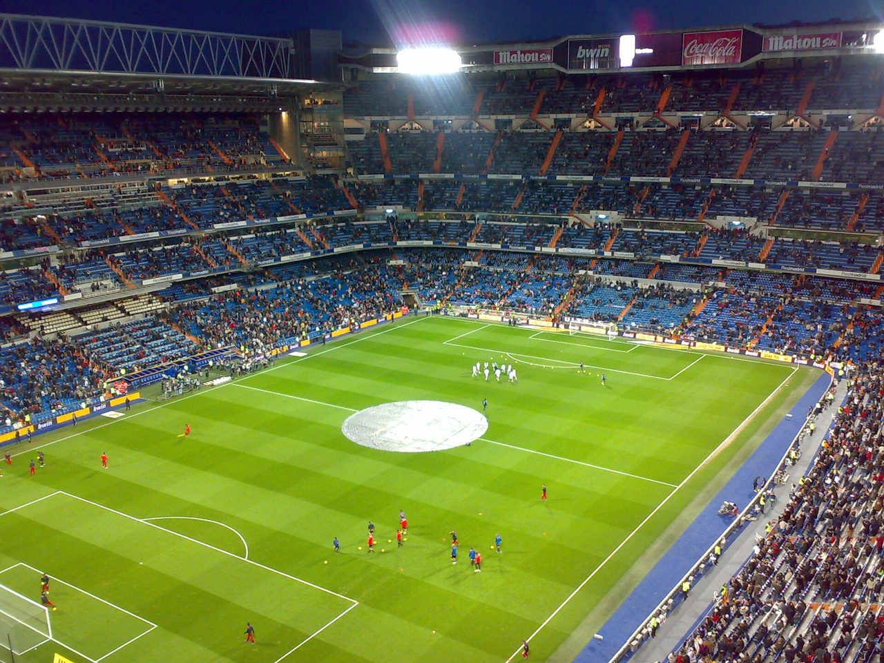 Interior del Santiago Bernabéu iluminado de noche, con el césped verde y los equipos calentando antes de un partido de Champions League