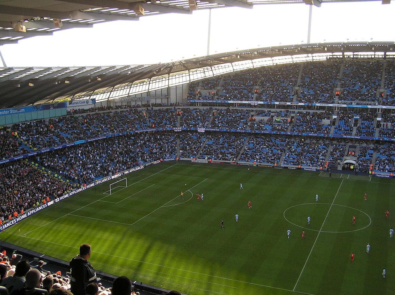 Partido de Premier League en curso en el Etihad Stadium, visto desde la tribuna alta con el césped rayado en primer plano